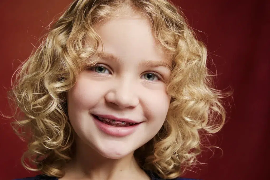 Young child with curly blonde hair smiling confidently with early orthodontic braces, reflecting positive early treatment results at Labbe Family Orthodontics.